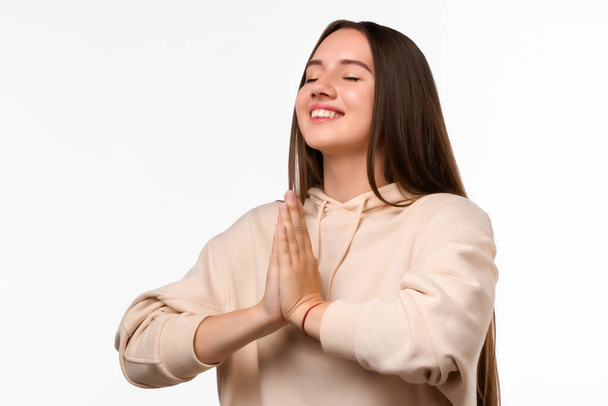 Image of pleased brunette woman with long hair smiling and holding palms together dressed in casual clothes. She meditating, praying for peace and love, having calm and peaceful facial expression - Fotografie, Obrázek