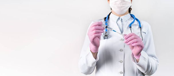 Обрезанный вид молодой женщины-врача в белом халате, в медицинской маске и розовых стерильных перчатках держит шприц. Сосредоточься на шприце. Концепция вакцинации. - Фото, изображение