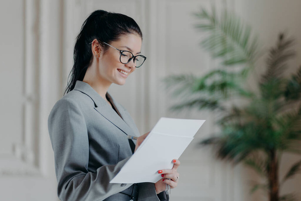 Glad female lawyer writes in corporate paper documents, wears optical glasses, formal clothes, stands in own cabinet, develops new strategy, has happy expression. People, career, employment concept - Photo, Image