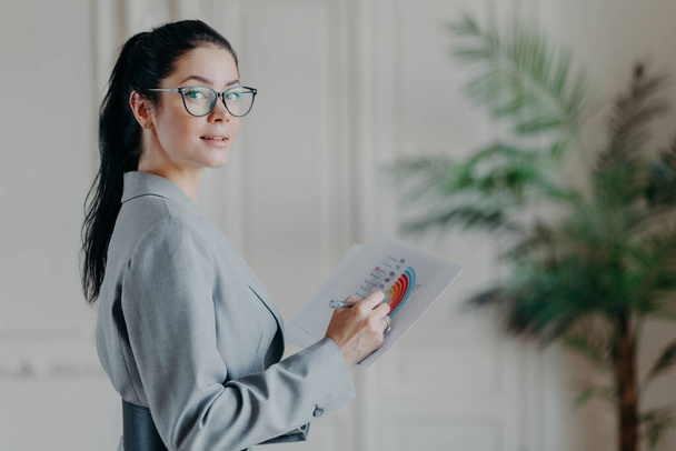 Horizontal shot of female strartuper analyzes business information, holds paper documents with diagrams and graphics, looks confidently at camera, wears transparent glasses and formal clothes - Фото, изображение