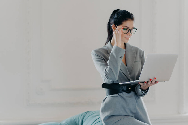 Елегантна професійна жінка-банкір, зосереджена на екрані ноутбука, одягнена в офіційний одяг, читає документи в онлайн-версії посад у просторій кімнаті, домашньому офісі. Бізнес, концепція технології
 - Фото, зображення