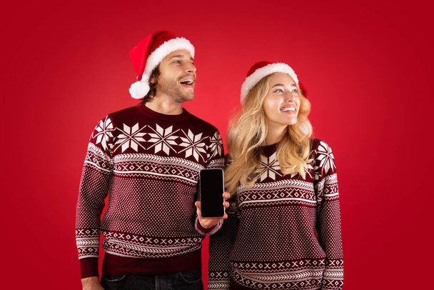 Joyeux couple millénaire en pulls de Noël et chapeaux de Père Noël regardant de côté et montrant téléphone portable avec écran blanc - Photo, image
