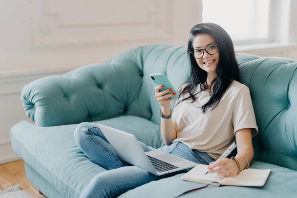 Glad female student works on course paper, surrounded with modern gadget, makes notes in notepad, feels relaxed at comfortable sofa, works online, wears casual clothes, watches video online. - Photo, image