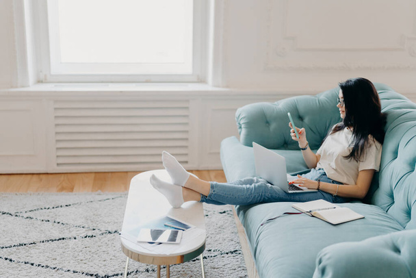 Sideways shot of relaxed female student uses modern technologies while doing homework, uses web sources for making project, sits on comfortable sofa against cozy interior, leans legs on desktop - Photo, Image