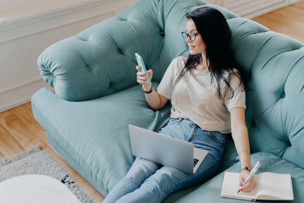 Photo of busy female freelancer in casual clothes focused in smartphone display, checks information in internet, notes something in diary, uses modern technologies for work, poses at cozy room - Foto, Bild