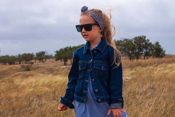 デニムジャケットの愛らしい女の子、黒サングラス、黄色の芝生のフィールドで青のプレイドレス。田舎の風景の上に幸せなスタイリッシュな長いブロンドの髪の子供。かわいい子供屋外田舎道の旅を歩く. - 写真・画像