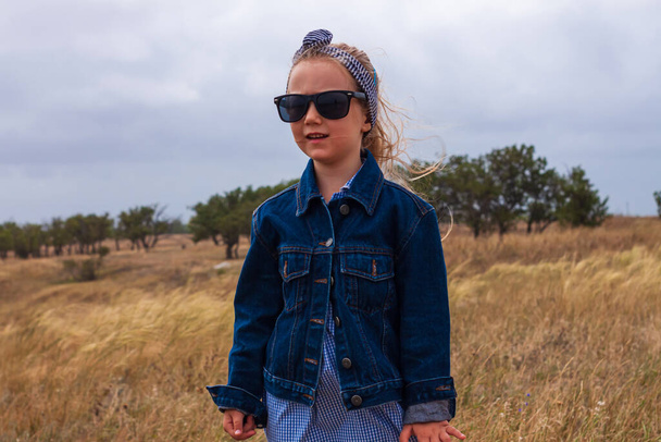 Adorable little girl in denim jacket, black sunglasses, blue plaid dress in yellow grass field. Happy stylish long blonde hair child on countryside landscape. Cute kid walking outdoor rural road trip. - Photo, Image