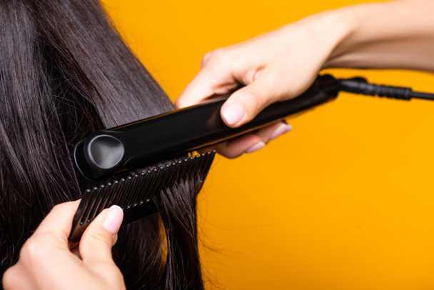 cropped view of hairdresser using hair straightener and comb isolated on yellow - Foto, immagini