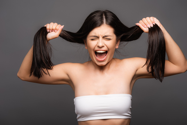 засмучена брюнетка жінка тримає пошкоджене волосся і кричить ізольовано на чорному
 - Фото, зображення