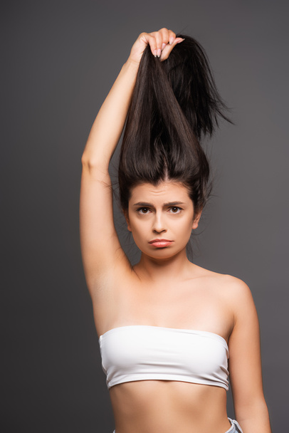 rozrušená bruneta žena drží poškozené vlasy izolované na černé - Fotografie, Obrázek