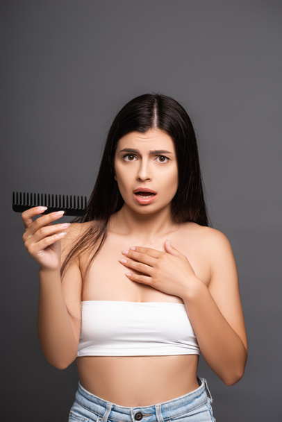 shocked brunette woman holding comb isolated on black - 写真・画像