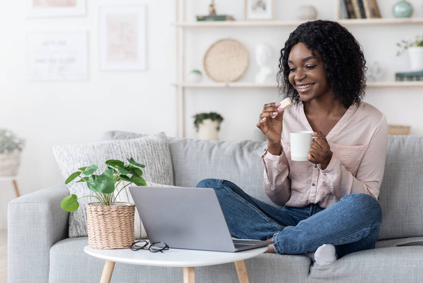 幸せなアフリカの女性はノートパソコンでコックでリラックス,お茶とワッフル - 写真・画像