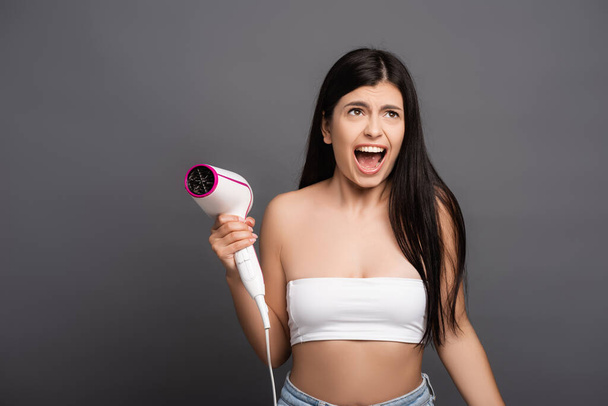 brunette woman holding hair dryer and yelling isolated on black - Foto, imagen