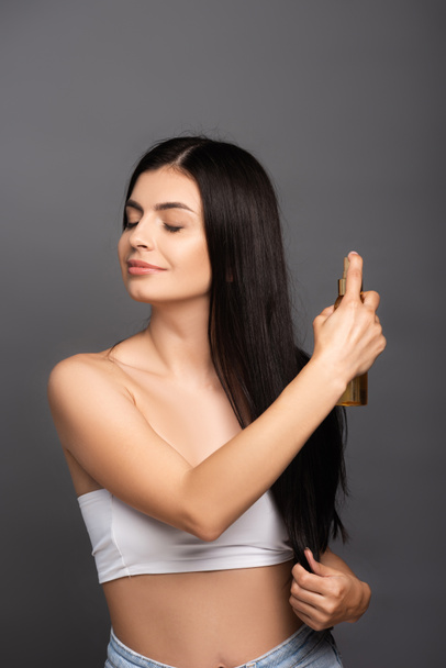 brunette woman spraying oil on hair isolated on black - Фото, зображення