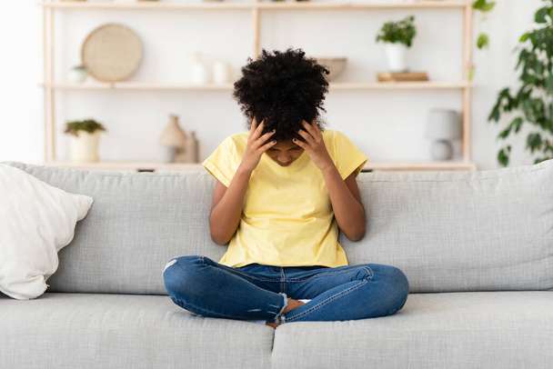 infeliz negra chica tocando la cabeza sentado en sofá en casa - Foto, imagen