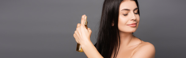 nahý bruneta žena sprejování olej na vlasy izolované na černé, panoramatický výstřel - Fotografie, Obrázek