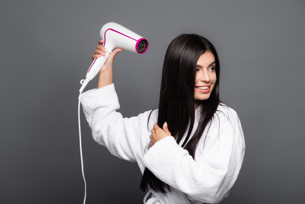 brunette long haired woman in bathrobe using hairdryer isolated on black - Foto, afbeelding