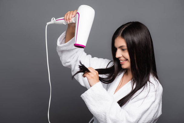 brunette long haired woman in bathrobe using hairdryer isolated on black - Фото, изображение