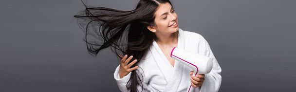 brunette long haired woman in bathrobe using hairdryer isolated on black, panoramic shot - Photo, Image