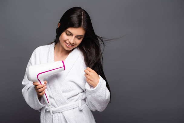 brunette long haired woman in bathrobe using hairdryer isolated on black - Photo, Image