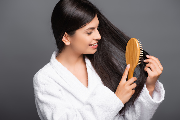 bruneta žena v županu kartáčování vlasy a úsměv izolované na černé - Fotografie, Obrázek