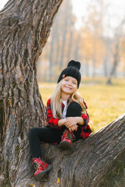 Пятилетняя девочка улыбается, сидя на дереве в осеннем парке в шляпе - Фото, изображение