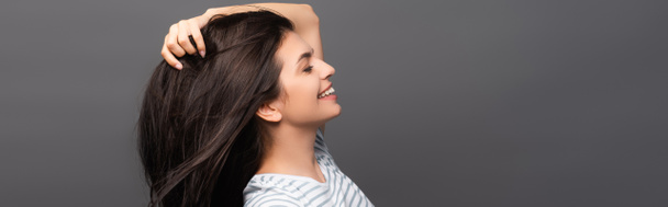 side view of brunette woman with closed eyes smiling and touching hair isolated on black, panoramic shot - Photo, Image