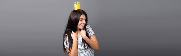 brunette long haired woman in paper crown with closed eyes smiling isolated on black, panoramic shot - Photo, Image