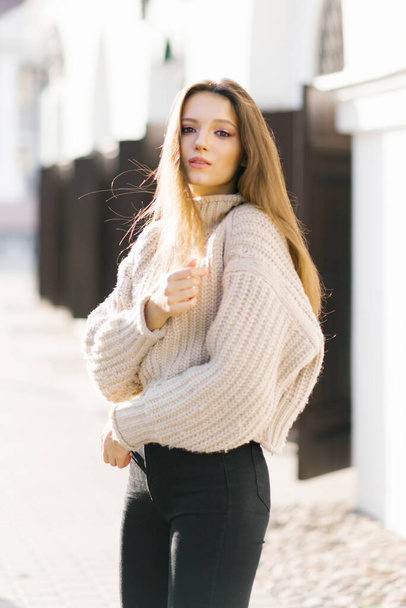 Porträt eines jungen schönen modischen Mädchens in einem beigen Strickpullover, das auf der Straße einer europäischen Stadt posiert. - Foto, Bild
