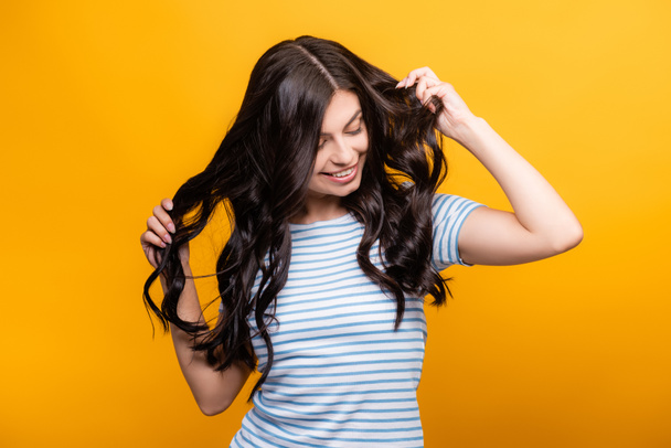 brunette woman toughing curly long hair and smiling isolated on yellow - Photo, Image