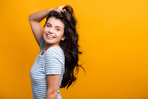 side view of brunette woman toughing curly long hair and smiling isolated on yellow - Fotoğraf, Görsel