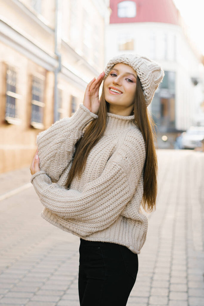 Autumn portrait: a young woman, dressed in a warm wool sweater and hat, poses on a city street and smiles - Foto, afbeelding