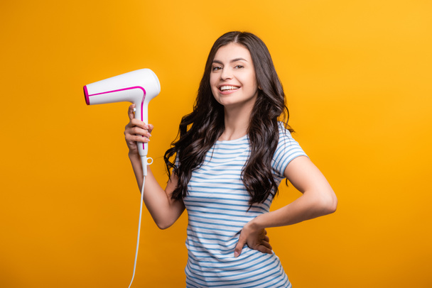 brunette woman with curls holding hairdryer isolated on yellow - Foto, Imagen