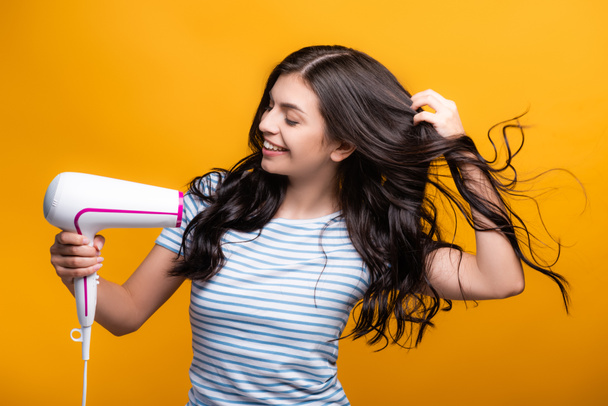 brunette woman with curls using hairdryer isolated on yellow - Photo, Image