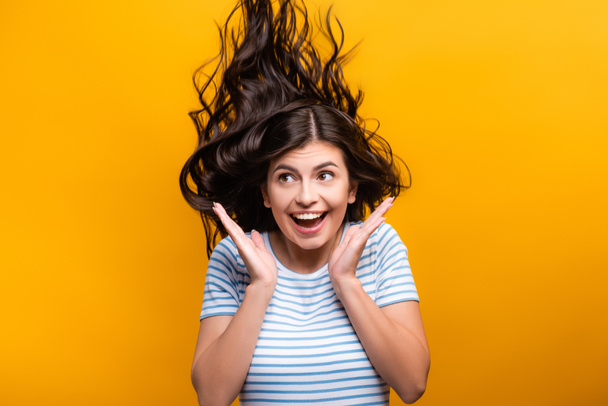 brunette long haired woman with curls jumping and smiling isolated on yellow - Фото, изображение