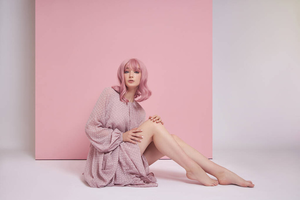A nő festett rózsaszín hajjal, hosszú ruhában a padlón ül. Portré egy lányról, akinek hajszíne van a rózsaszín falnál. Tökéletes frizura és frizura - Fotó, kép