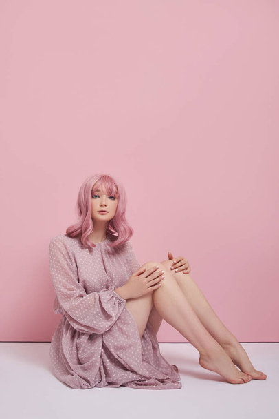 Woman with dyed pink hair in a long dress is sitting on the floor. Portrait of a girl with hair coloring at the pink wall. Perfect hairstyle and hair styling - Photo, Image