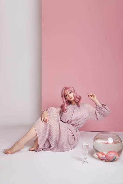 Girl with dyed pink hair in long dress sitting on the floor with a round aquarium. Portrait woman with hair coloring at pink wall. Perfect hairstyle and hair styling - Photo, image