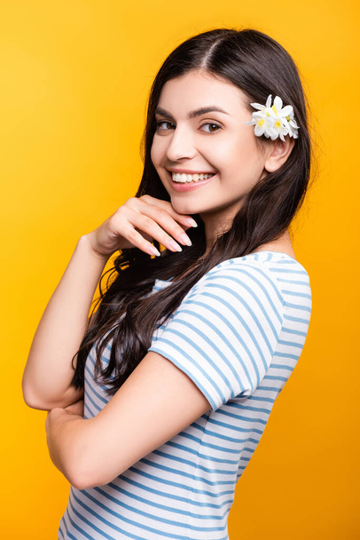 μελαχρινή νεαρή γυναίκα με λουλούδια στα μαλλιά χαμογελώντας απομονωμένη στο κίτρινο - Φωτογραφία, εικόνα