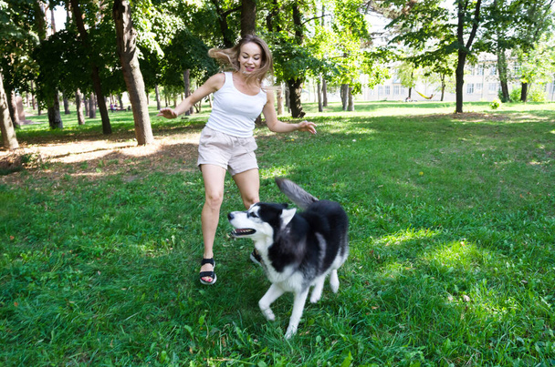 atrakcyjna blondynka biegnie i bawi się z psem w parku. - Zdjęcie, obraz