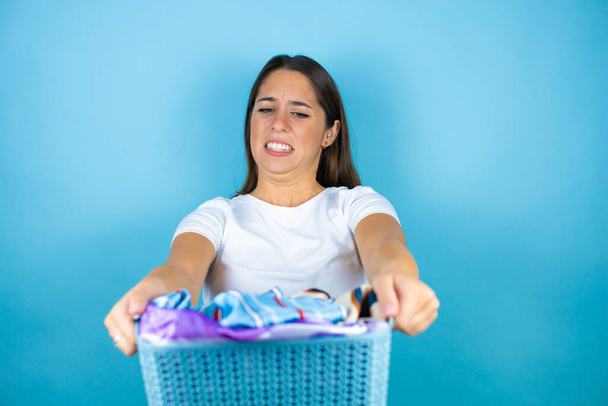 Joven hermosa mujer sobre fondo azul aislado haciendo tareas domésticas sosteniendo una canasta maloliente con ropa - Foto, imagen