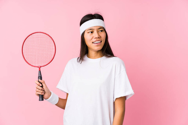 Pembe arka planda badminton oynayan genç Çinli kadın gülümseyen, neşeli ve keyifli bir kenara bakar.. - Fotoğraf, Görsel