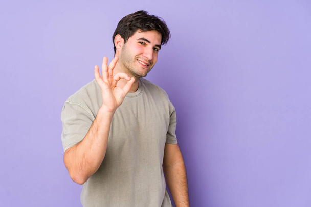Junger Mann auf violettem Hintergrund zwinkert und hält eine Okay-Geste mit der Hand. - Foto, Bild