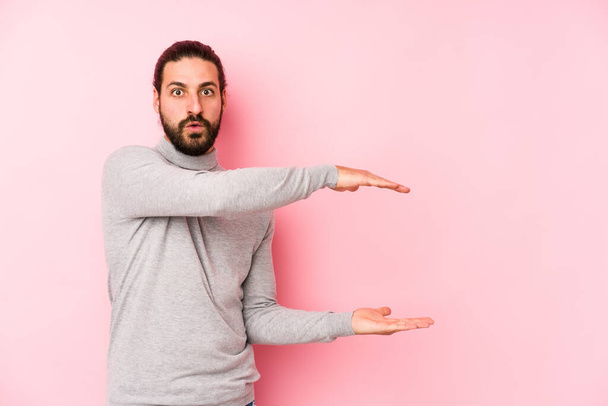 Young long hair man isolated on a pink background shocked and amazed holding a copy space between hands. - Photo, image