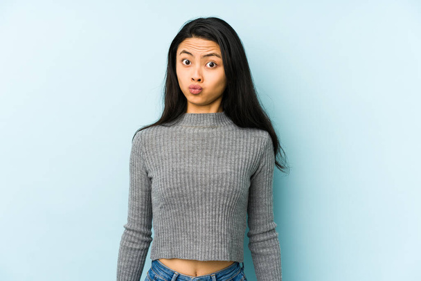 Young chinese woman isolated on a blue background blows cheeks, has tired expression. Facial expression concept. - Photo, Image