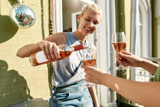 Cheerful girl in casual wear pouring wine while standing on the balcony together with her girlfriend. Young lesbian couple spending their day together - Photo, image