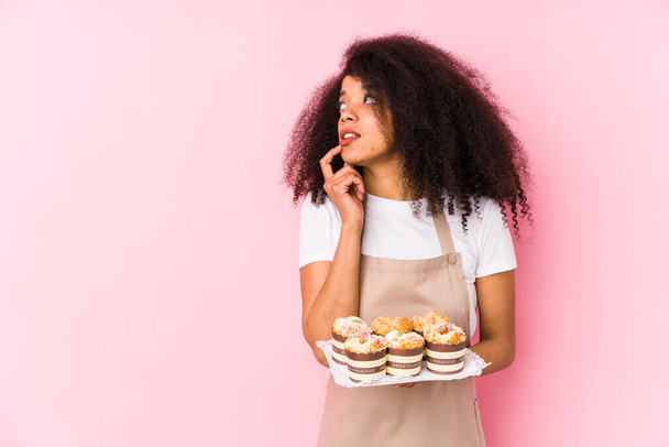 Mladý afro cukrář žena drží cupcakes izolovanýMladý afro pekař žena uvolněně přemýšlet o něco při pohledu na kopírovací prostor. - Fotografie, Obrázek