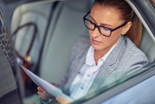 Konzentrierte Geschäftsfrau mittleren Alters mit Brille, die Dokumente liest, während sie auf dem Rücksitz im Auto sitzt und sich auf ein Treffen vorbereitet - Foto, Bild