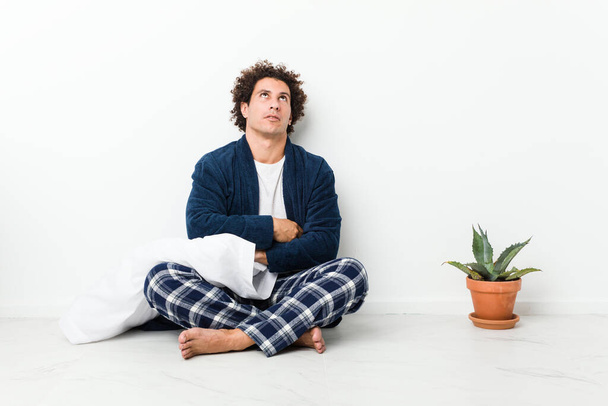 Dojrzały mężczyzna w piżamie siedzi na podłodze domu zmęczony powtarzającym się zadaniem. - Zdjęcie, obraz