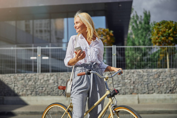 Χαρούμενη χαρούμενη γυναίκα με λευκό κινητό τηλέφωνο στο χέρι ξοδεύοντας χρόνο στο άδειο δρόμο της πόλης - Φωτογραφία, εικόνα